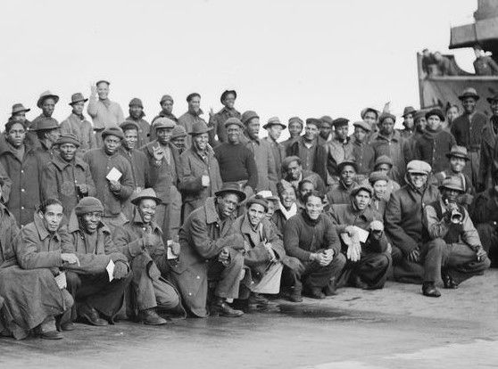 A group of 50-60 men pictured, standing and kneeling on the quayside in Greenock, Scotland in November 1942.  All are black and are from British Honduras (now Belize), having travelled to do wartime forestry work.  The men are wrapped up in what seem to be somewhat unfamiliar layers of warm clothes, but most are smiling broadly and some are giving the thumbs up.  Almost all are wearing hats, and one man kneeling and wearing a trilby is ready to play the trumpet that he has brought with him.  