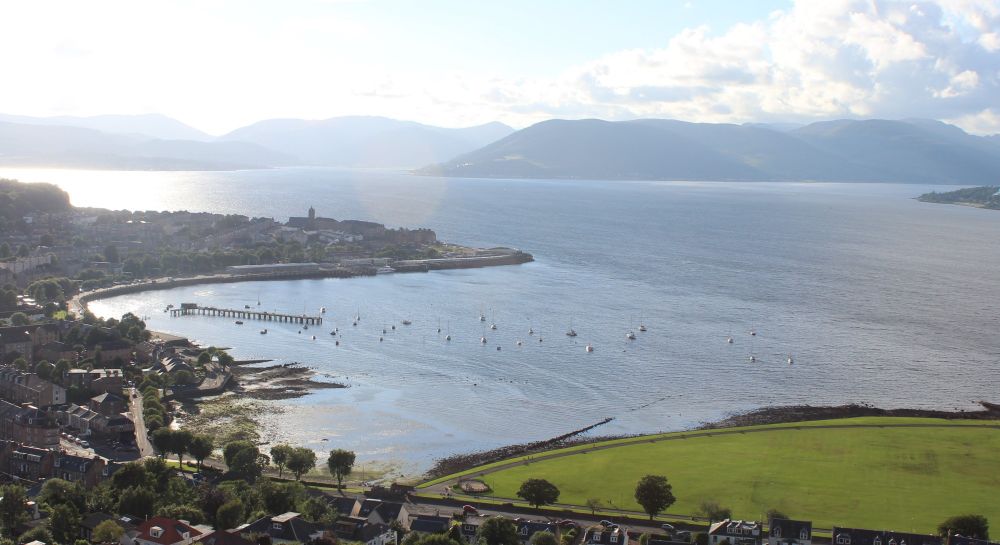 From Lyle Hill looking north over the Firth of Clyde, in 2024.  No ships are visible.  The landscape is the same as the 1945 picture.  The town of Gourock appears to stick out into the waters of the Clyde.  Clouds, and the hills of Argyle and Bute, loom beyond.  Picture is in colour. 