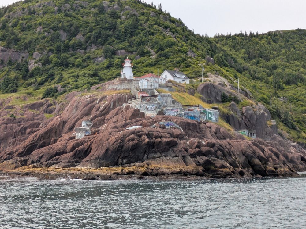 Fort Amherst, viewed from the water looking up.  The fort is situated above 40 feet of rocky cliffs.  1940s concrete buildings are constructed on areas that have clearly been flattened out from the cliffs for the purpose of construction.  At the top, a lighthouse, still in operation, guides shipping.   Above, thin foliage covers the rocky hills.  