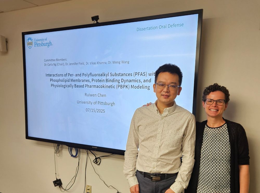 Newly minted PhD and proud Professor stand in front of PowerPoint slide on screen displaying title of dissertation: "Interactions of PFAS with phospholipid membranes, protein binding dynamics, and PBPK modeling"