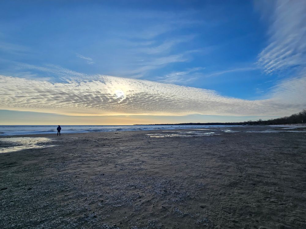 A long stretch of sand reaches towards the horizon where snow and frozen lake meet beneath deep blue sky and a low evening sun hides behind a striated bank of luminescent clouds. Left of center the dark outline of a solitary figure stands framed between sand and sky.