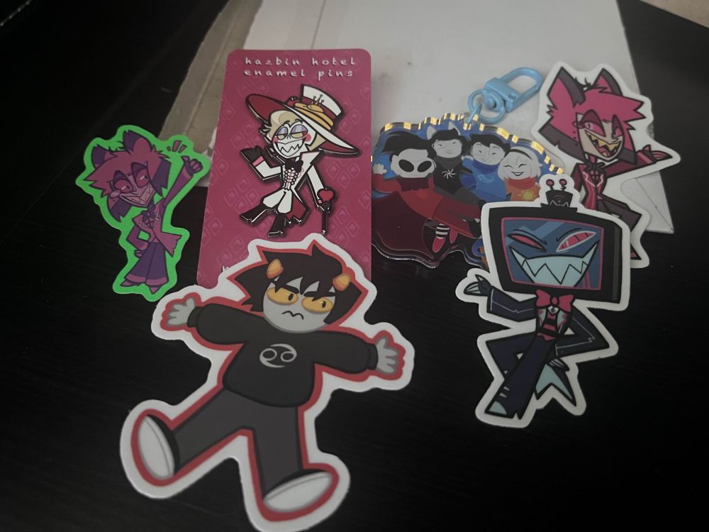 Left to right - Sticker of Alastor, pin of Lucifer, sticker of Karkat Vantas, charm of the beta kids, sticker of Vox, different sticker of alastor