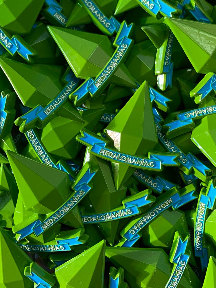 Photo of an enamel pin shaped like a Sims plumbob with a banner that reads “megalomaniac”