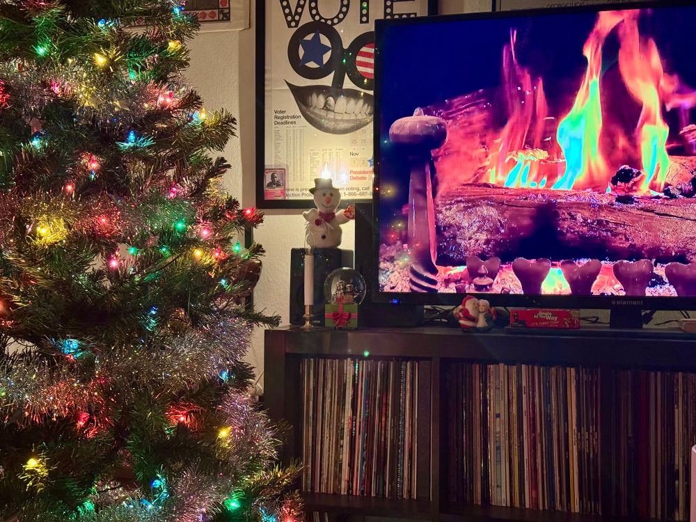 Photo of a Christmas tree with incandescent lights shining next to a full record shelf and a tv above with a fireplace on it— a cozy Christmas scene is set