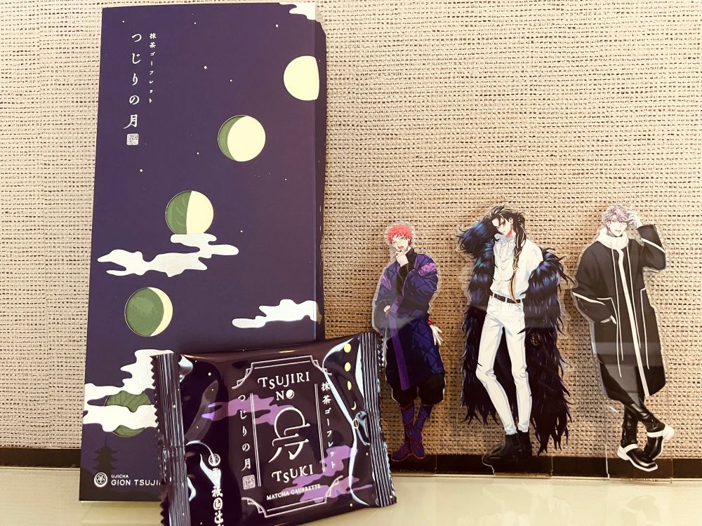 左側紫色の箱に満ちていく月の絵が描かれた菓子箱と、その個包装（紫色の袋の中央に月の絵と漢字を組み合わせた意匠、右側に小さく満ちていく月の絵）
その横にEW04衣装のB.A.T.アクスタが左から番手順に並んで立っている
