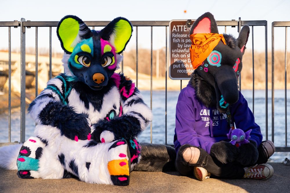 Flicker (African Wild Dog) and Matrix (the creature) sit calmly cross-legged in front of a metal railing. A dam and river are in the background 