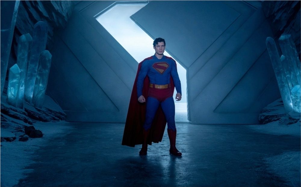 Superman stands in his fortress of solitude wearing his iconic red cape and blue outfit with a red and gold logo on his chest. Scene from the 2025 comic book adaptation by James Gunn.
