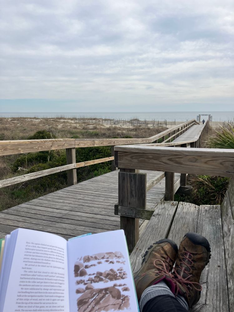 Open book and boots showing as person reads the book with a pier stretching out toward the ocean. The ocean and sky are shades of light blue and white. 