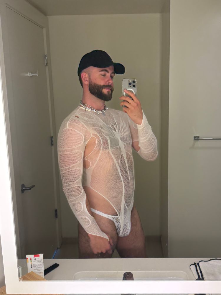 person wearing a white mesh bodysuit and a white thong taking a mirror selfie in a bathroom at a front-facing 3/4 angle.