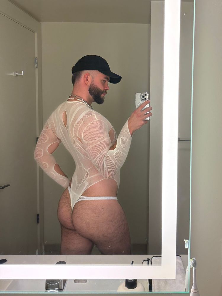 person wearing a white mesh bodysuit and a white thong taking a mirror selfie in a bathroom at a back-facing 3/4 angle.