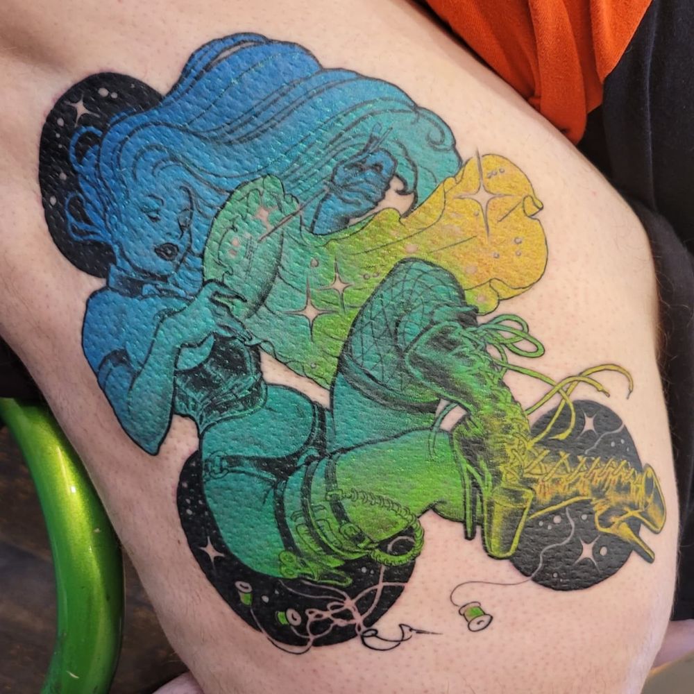 A closer shot of the completed tattoo, it is a bit swollen but you can see a feminine person laying down. She is wearing an underbust corset that stops above her belly button, a strap on harness, leg harnesses with a scissor holster, a bunch of stitching needles and a fishnet stocking on the other leg. She has a collar on and some super high heeled goth boots. Her long hair is flowing up around her face and behind the cross stitch hoop in her hands. The figure is holding the cross stitch hoop in one hand and pulling the needle and floss through with the other. The figure is coloured in a gradient going from blue to teal to green to yellow, and the cross stitch fabric is a teal/green to a yellow in lighter shades. This one is a mid-zoom shot so you can also see the three black circles at her head, ass and feet that make up the background. These sections have some negative space stars and sparkles, as well as negative spaces floss strands from green spools of floss