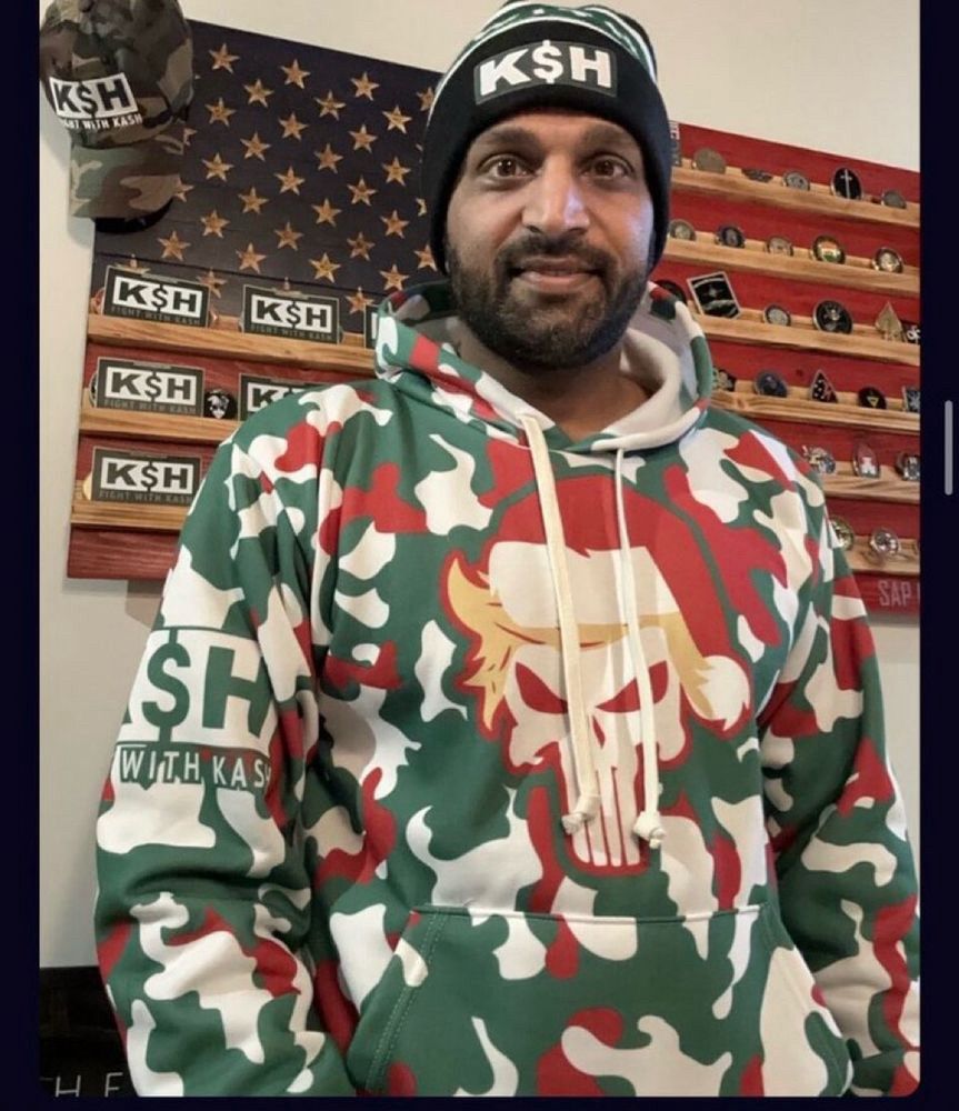 Kash Patel wearing a psychotic red and green camo hoodie featuring the Punisher with Trump’s ridiculous constructed hair and a hat that says K$H. He looks completely idiotic. 