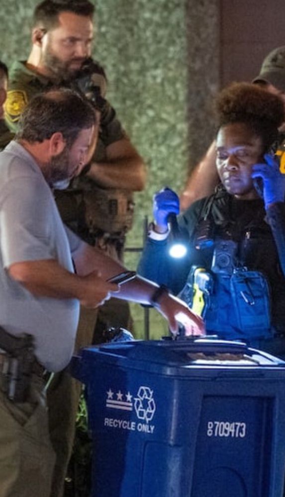 Zoom in on the photo to see a cop with a gut taking a cell phone photo of something in a recycling bin next to what looks like a science tech in gloves with a flashlight pointing inside the recycling bin. They look very serious. 