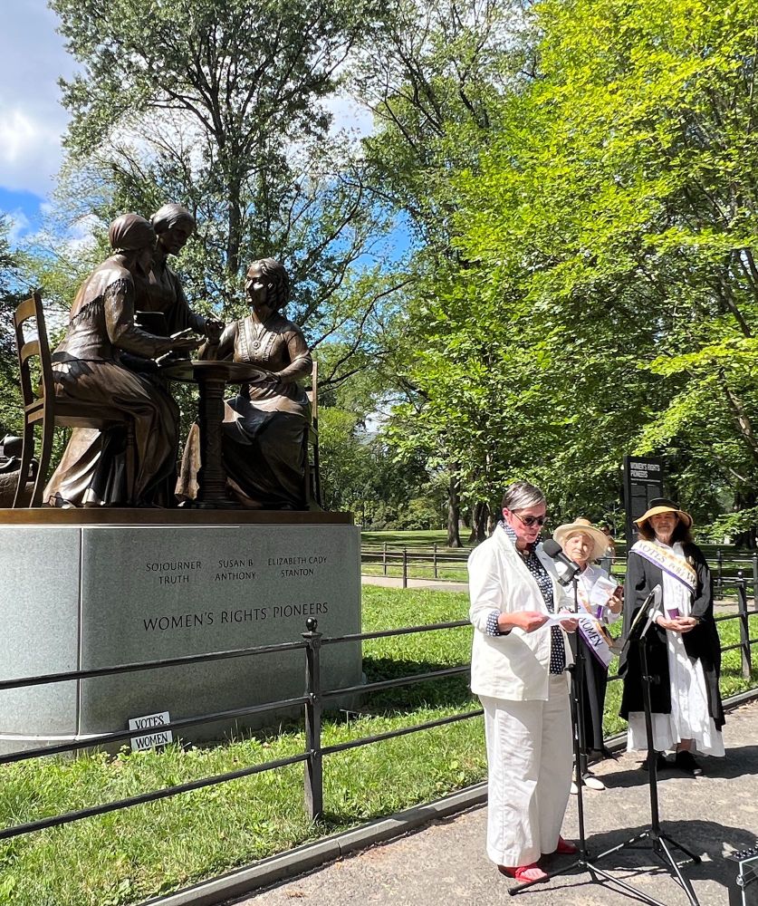 Three women, two of them wearing suffragette sashes, one reading a speech, standing in front of a fairly new statue in Central Park depicting three women's rights pioneers. 