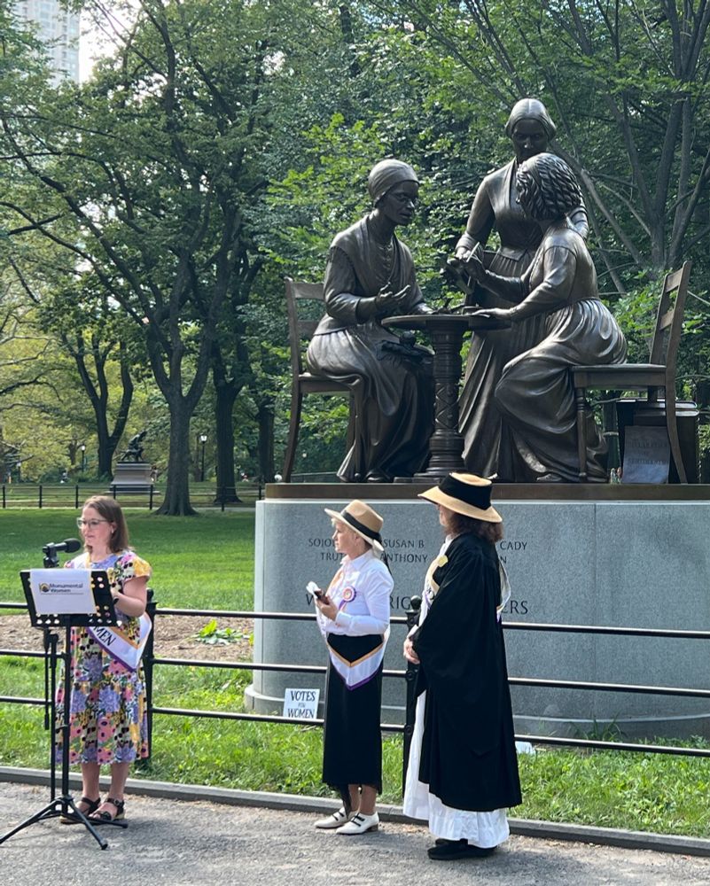Three women, all wearing suffragette sashes, one reading a speech, standing in front of a fairly new statue in Central Park depicting three women's rights pioneers. 