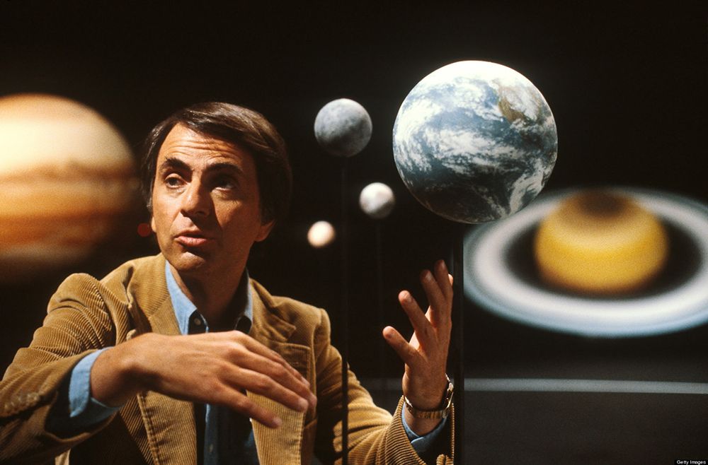 Carl Sagan gesturing in front of models of planets