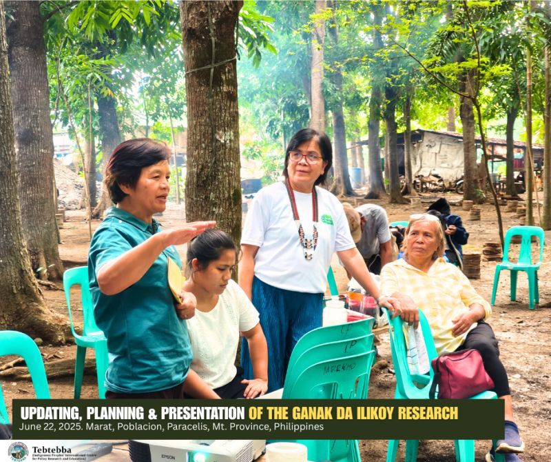 Picture from Tebtebba a group of women are standing or sitting in teal plastic chairs outside in nature surrounded by trees.  One is explaining something while the others listen  