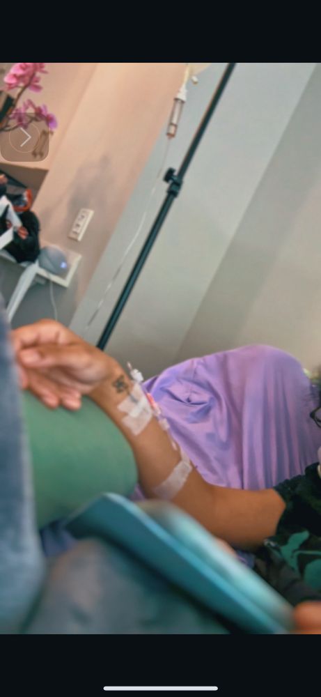 Brown skin woman on iPad in bed. There’s an iv pole in the background and tubes connected to her arm 