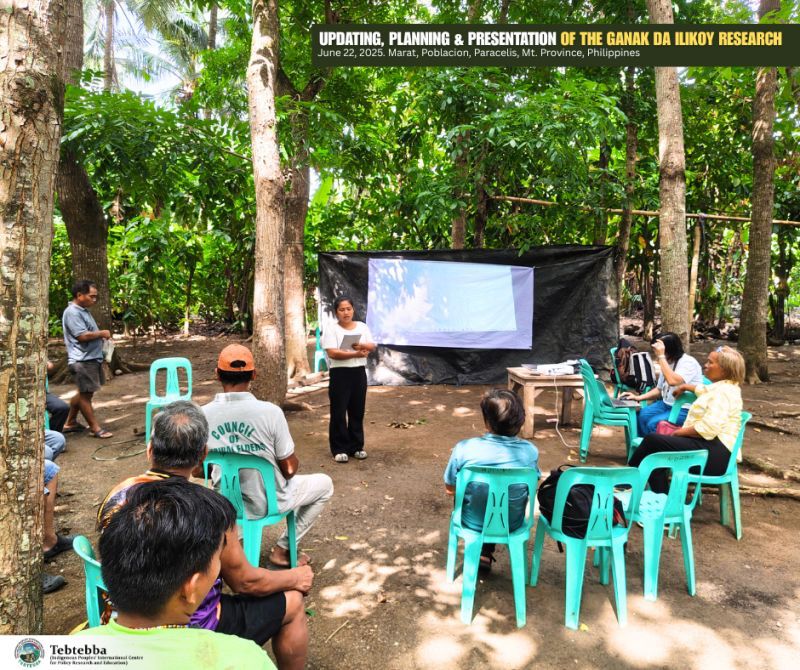 There’s a group of people sitting around in teal plastic chairs watching a presentation on a black sheet hanging up with a screen being projected onto it. The sheet is tied to two trees
