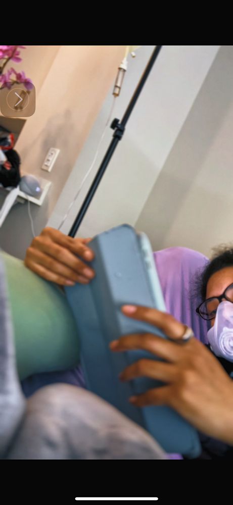 Brown skin woman on iPad in bed. There’s an iv pole in the background 