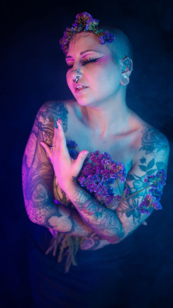 Arachnie grips purple flowers in her arms, with matching flowers resting on needles pierced into her forehead. 