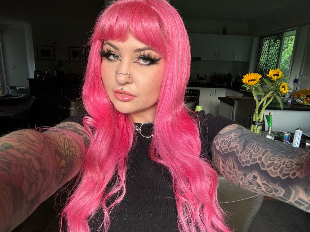 An arm’s length selfie of Arachnie. She has two full-sleeve arm tattoos and long pink hair. Sunflowers are in a vase behind her 