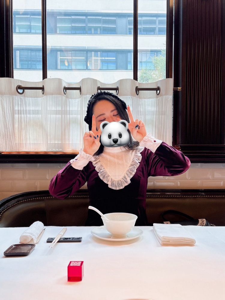 食器がセットされた白いクロスの引かれた大きなテーブルについて、両手でピースサインをしている同行者の写真。ヨーク部分にレースフリルのあしらわれたボルドーのベロアのワンピースを着ており、ベロア素材の黒いヘッドドレスをつけている。