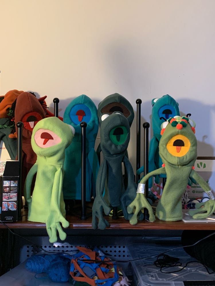 several in-progress fleece puppets, some with no eyes and some with blank white eyes with no pupils, they are all hanging on a rack so their mouths are wide open