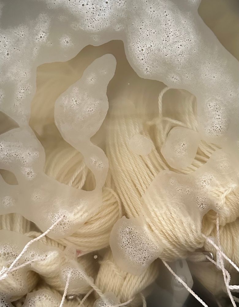 Many skeins of white wool yarns tied with baker’s twine, sitting in a soapy bath in a large stainless steel pot 