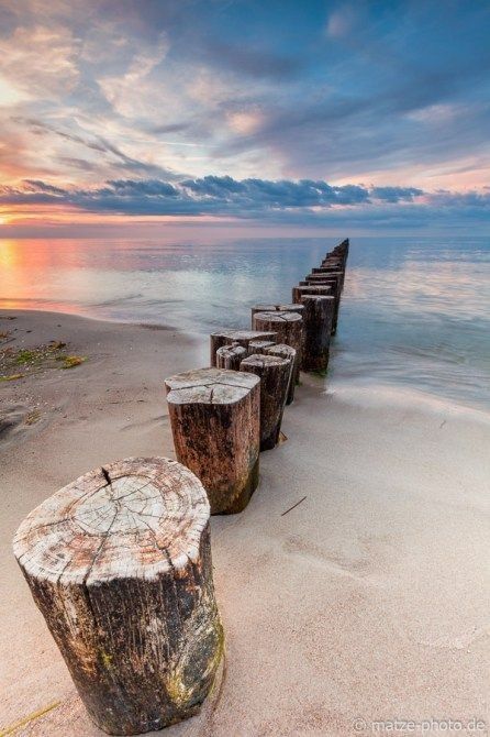 Im Vordergrund stehen dicke Holzfäller, die aufgereiht vom Strand in Meer Verlauf. Das Wasser und die Wolken sind von der untergehenden Sonne in Pastellfarben gefärbt. 

Foto/ Pixabay 