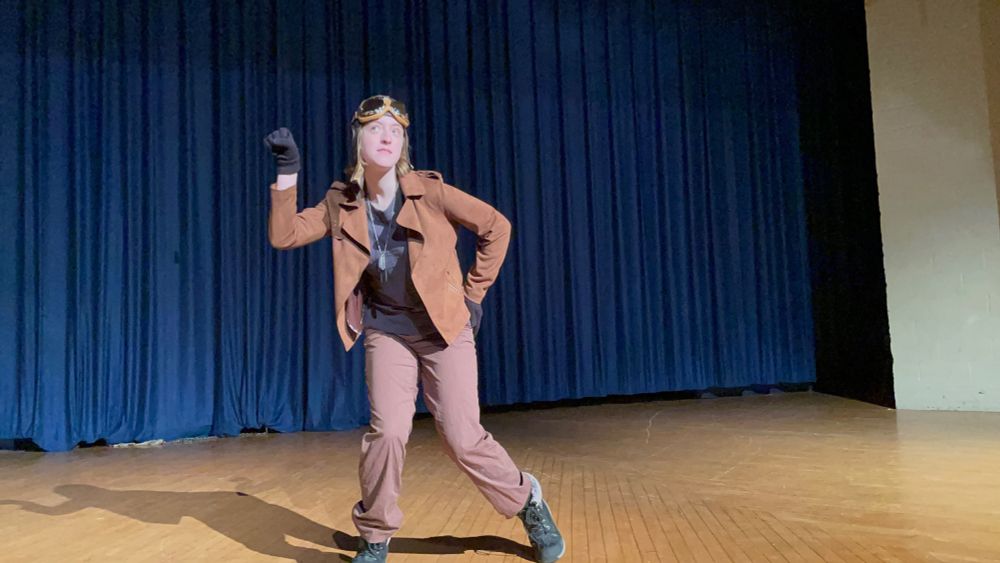 (1) A photo of Jet Set Jackie (myself) doing a drag set to "Airheart" by Steam Powered Giraffe. I am wearing a brown jacket and pants, black boots, and an old fashioned aviator's helmet and goggles.