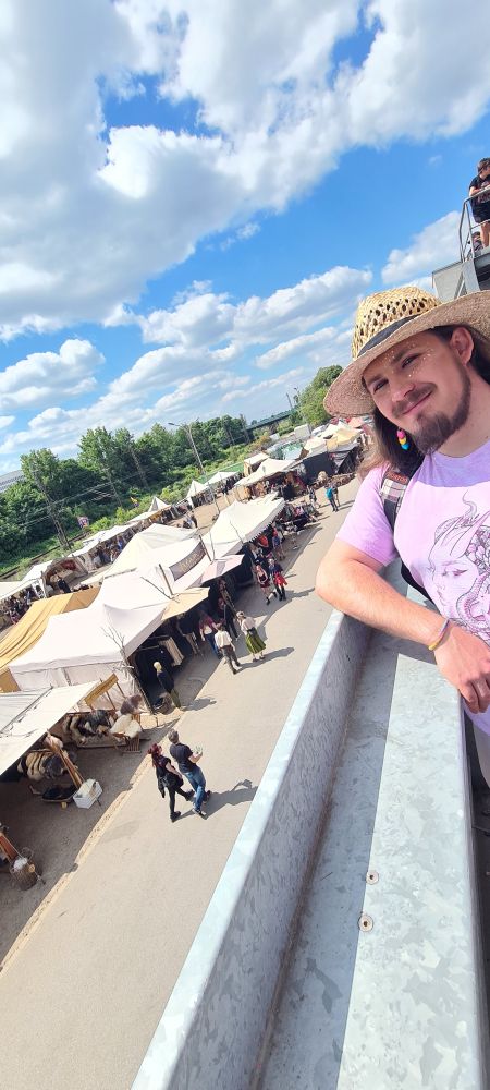 An elevated view of a medieval market. Many tents stand on concrete ground with people walking before them. The photo is taken from a balcony above and with an angle, so another person leaning on the balcony is visible. The person smiles, wearing a strawhat and a pink shirt with open brown, long hair, a beard and an earring with the pan flag colours. 