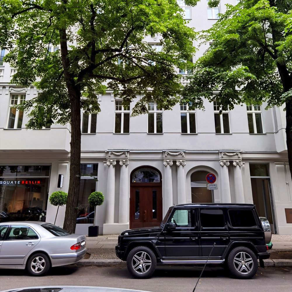 Das Foto zeigt das mehrgeschossige, alte Mietshaus in der Schlüterstraße 45 in Berlin-Charlottenburg. Das Haus ist ein renovierter Altbau mit heller, sauberer Fassade und zahlreichen schmalen Fenstern. Der Blick auf dem Bild führt über die Straße und hindurch zwischen zwei Pkw, die vor dem Haus geparkt sind, sowie zwischen zwei großen, üppig grünenden Laubbäumen, die sich an den beiden Bildrändern befinden. Die Haustür der Schlüterstraße 45 besteht aus dunklem, stellenweise verglastem Holz und ist von Stuck umgeben, der antiken Säulen mit prachtvollen Kapitellen nachempfunden ist.