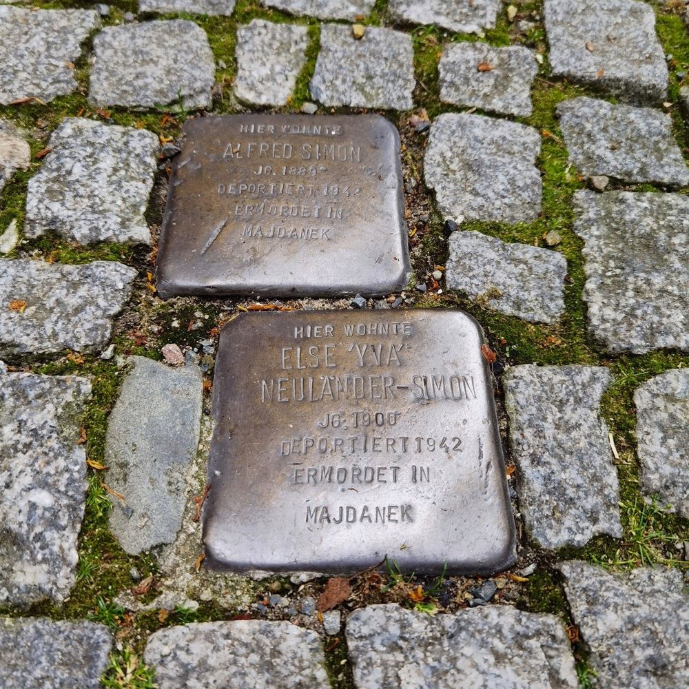 Das quadratische Foto zeigt die beiden Stolpersteine in der Schlüterstraße 45 in Berlin-Charlottenburg, darauf die folgenden Inschriften: "Hier wohnte Alfred Simon, Jg. 1889 - Deportiert 1942 - Ermordet in Majdanek" und "Hier wohnte Else 'Yva' Neuländer-Simon, Jg. 1900 - Deportiert 1942 - Ermordet in Majdanek". Der Stein zu Ehren von "Yva" befindet sich im Bildvordergrund, der Stein zu Ehren von Alfred Simon befindet sich (leicht nach links versetzt) dahinter. Die Stolpersteine sind von zahlreichen kleinen, grauen Pflastersteinen umgeben, zwischen denen sich grünes Moss befindet. Die Stolpersteine sind sauber, ihr Metall glänzt matt und leicht bräunlich.