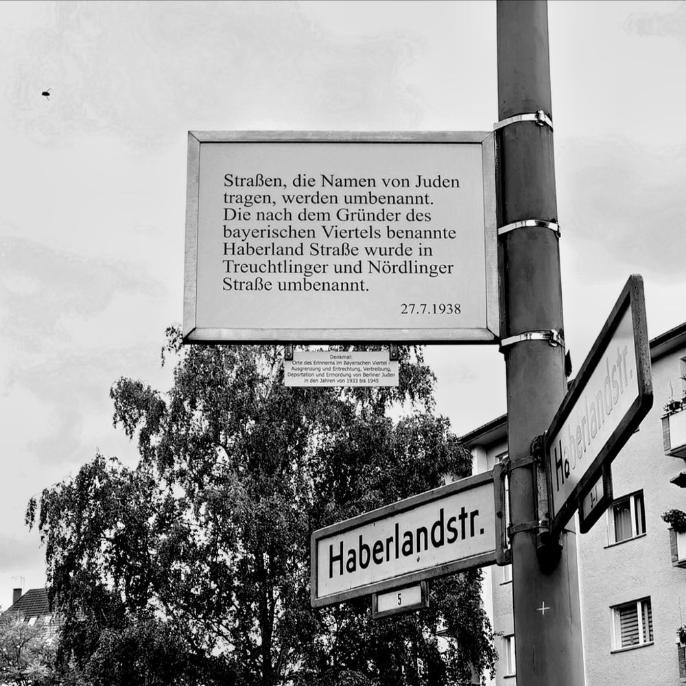 Das schwarz-weiße Foto zeigt eine Gedenktafel im "Bayerischen Viertel" in Berlin-Schöneberg, darauf die folgende Inschrift: "Straßen, die Namen von Juden tragen, werden umbenannt. Die nach dem Gründer des bayerischen Viertels benannte Haberland Straße wurde in Treuchtlinger und Nördlinger Straße umbenannt. 27. 7. 1938". Die Straße befindet sich an einem Laternenpfahl am rechten Bildrand und über zwei Straßenschildern der heutigen Haberlandstraße, schwarz beschriftet auf weißem Grund. Das Mahnzeichen hebt sich zudem von einer Baumkrone, einem mehrgeschossigen Mietshaus und einem dicht bewölkten, aber hellen Himmel im Bildhintergrund ab.