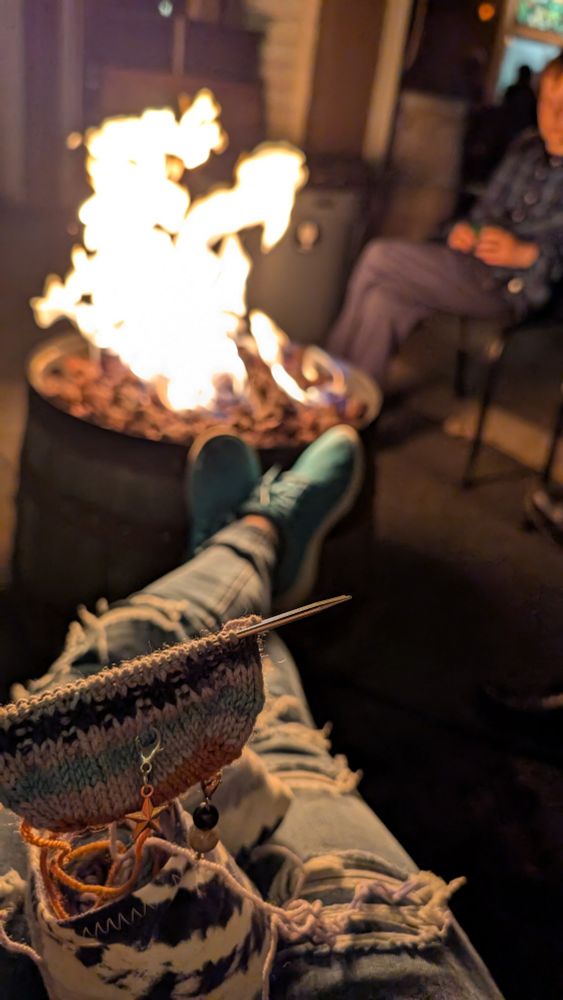 The toe of a new knit sock on chiaogoo red lace knitting needles with an outdoor fire pit in the background. 