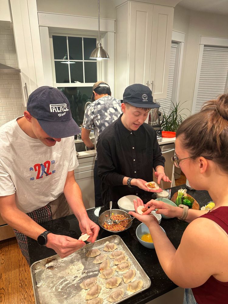 3 friends hand make pork and shrimp dumplings while one cleans dishes in the sink.