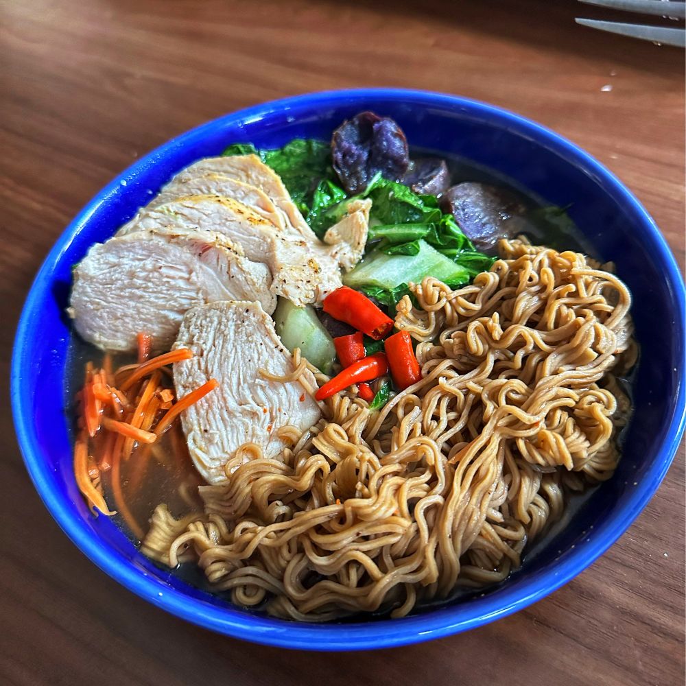 A azure bowl filled to the brim with ramen noodles, chicken fillet, red Thai chilies, purple potatoes, and a medley of greens in the dark broth.