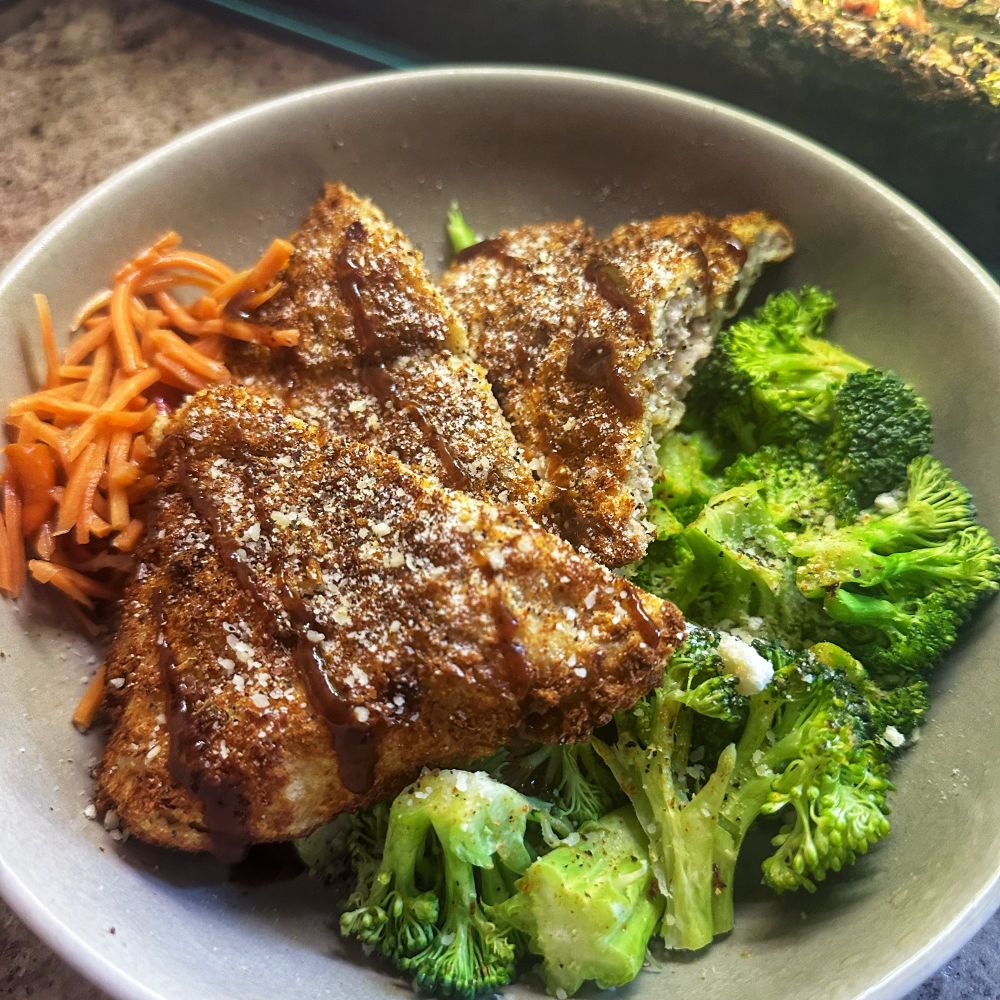 A cook meal in a grey stoneware bowl shot from above. Baked breaded chicken layered on top of a bed of seasoned broccoli, with orange carrots on the side. 