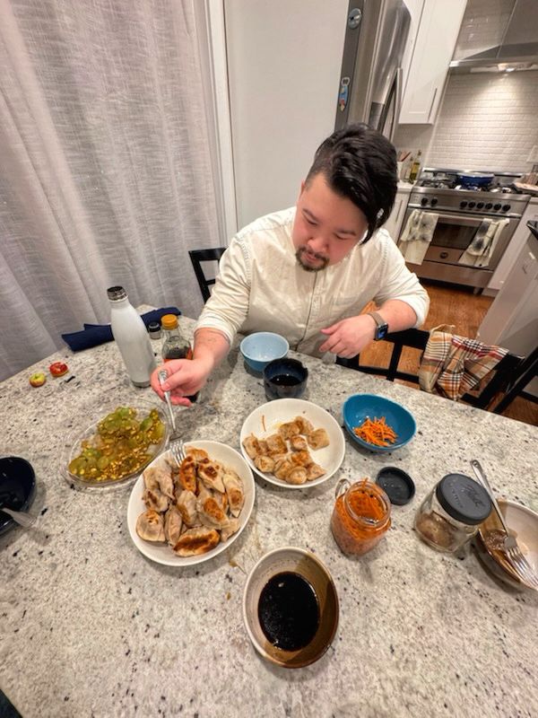 A picture of a man forking a dumpling out of a large plate of them. There is a large food spread before him which includes many dumplings, pickled carrots, veggie sides, and several dipping sauces. 