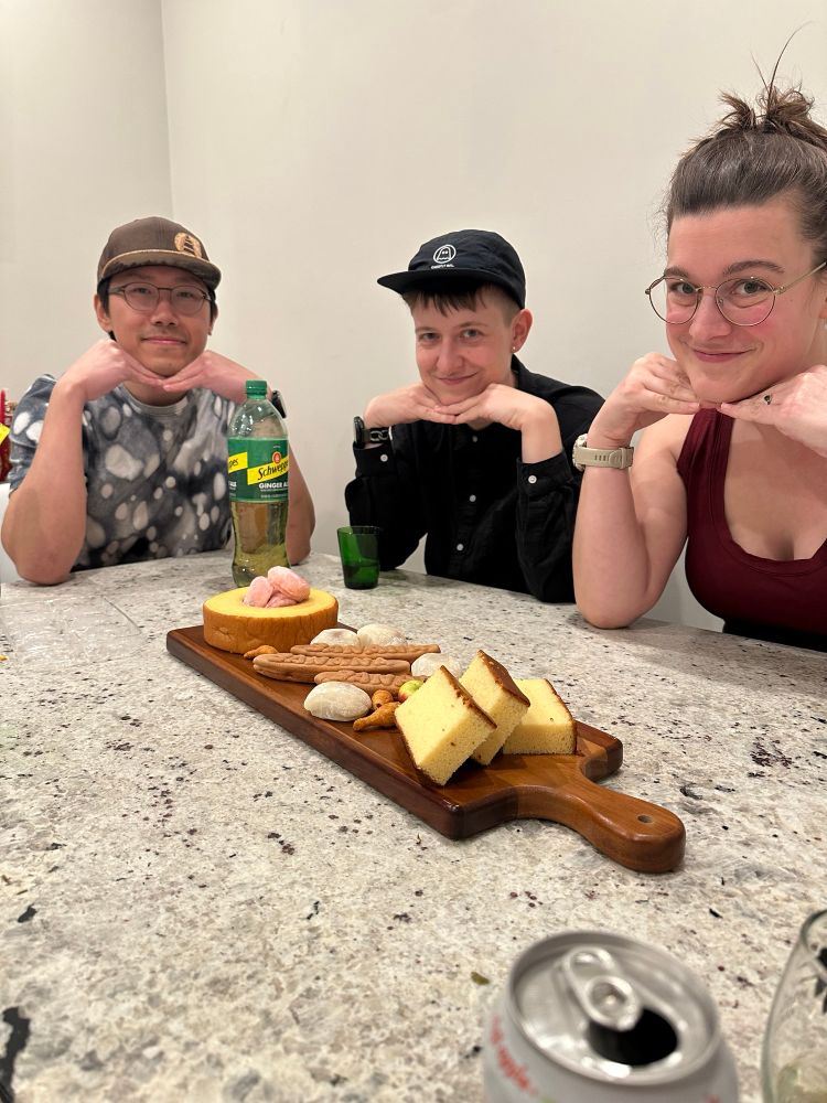 3 friends pose and smile for the camera with their elbows on the table and fingers tucked under the chin above a charcuterie board. The board contains several snacks and baked goods from HMart.