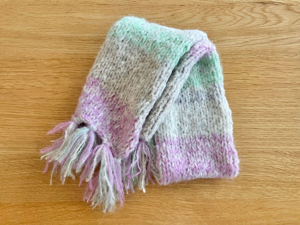 モヘヤ系の余り毛糸で編んだマフラーをラフに折りたたみ、木の机に置いて俯瞰で撮影した写真。マフラーは、白っぽい毛糸をベースに淡い紫、ミントグリーンを引き揃えてボーダー柄にしています。縁には本体を編んだのと同じ毛糸で作ったフリンジをつけてあります。