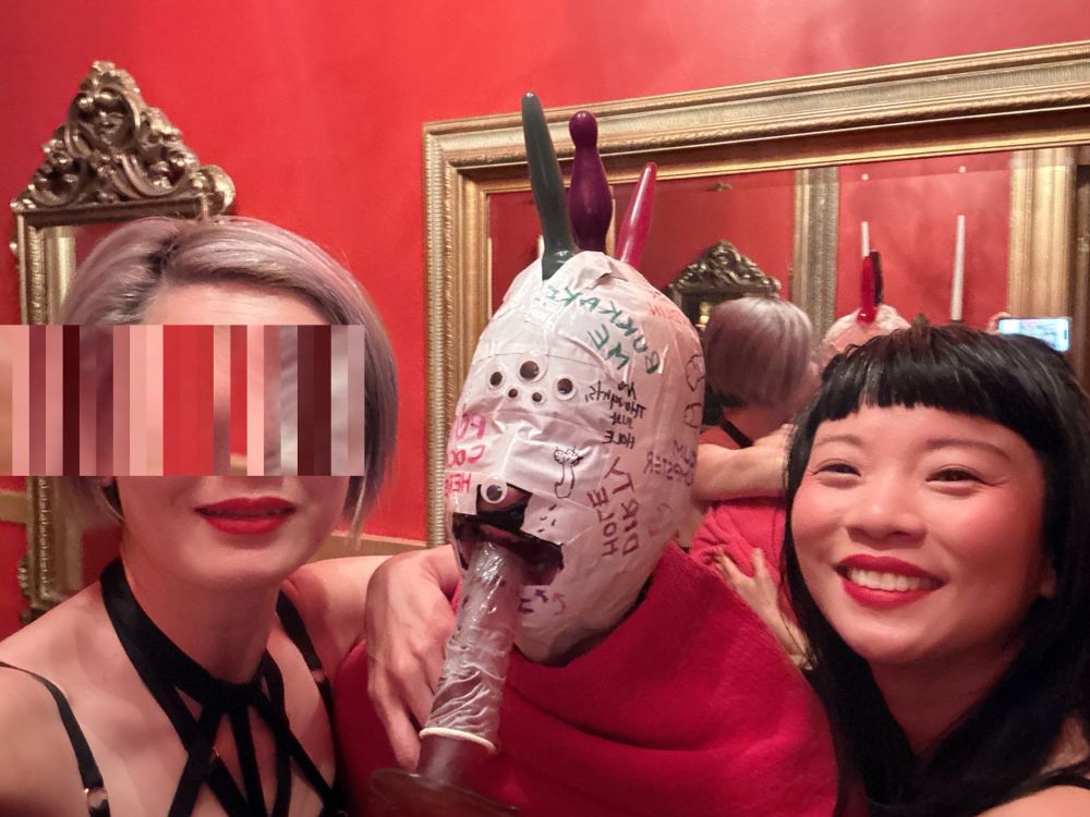 Domina Yuki and Domina Dia Dynasty posing and smiling with our creation- a glory hole goblin. He has a mohawk made from butt plugs, face wrapped in white tape with dirty messages written on it, multiple googly eyes and a dildo stuffed in his hole. 