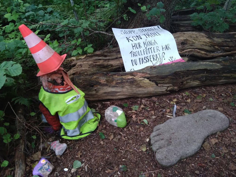 Ett troll med kon på huvudet. En stor betong fot. Skylt: "Stopp! Kon troll! Hur många trollfötter har du passerat?"