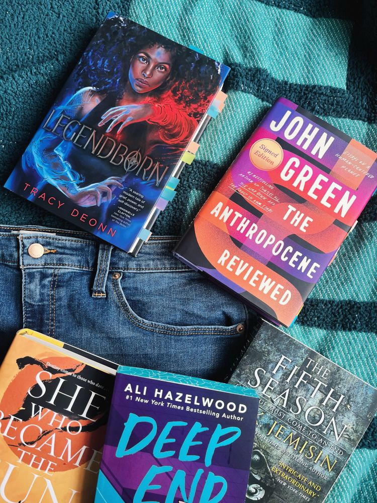 The picture is a pair of blue jeans with various books on one of the legs. The books include, from left to right and top to bottom, Legendborn, The Anthropocene Reviewed, She Who Became the Sun, Deep End, and The Fifth Season. 