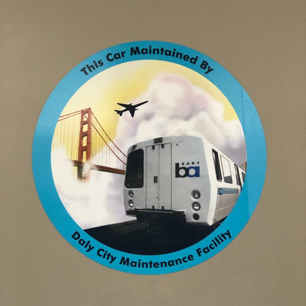 Roundel sticker depicting a BART train cutting through fog in front of the Golden Gate Bridge as an airplane soars above. The roundel says: “This Car Maintained by the Daly City Maintenance Facility”.