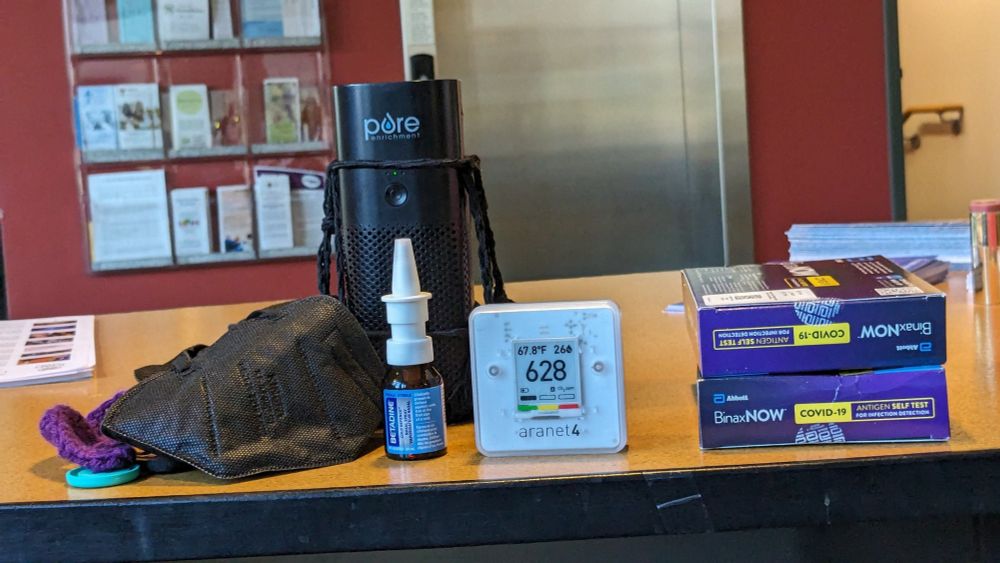 A collection of objects on a desk. From left: a KN95 mask with a handmade "ear saver"; a personal HEPA air filter; betadine nasal spray; an ARANET4 Carbon Dioxide monitor; and two boxes of BinaxNOW COVID Rapid tests