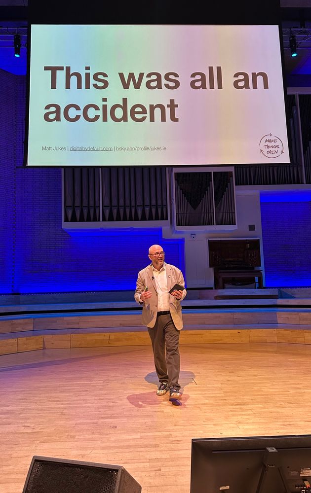 A white man on stage in front of a large screen that says “This was all an accident”