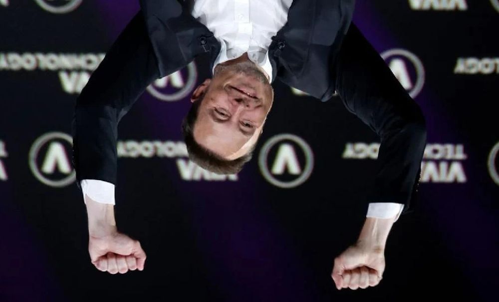 elon musk de ponta cabeça (a pose original é ele levantando os braços para comemorar algo, mas ela foi virada de forma a parecer que ele está pendurado e com os braços em suspensão)