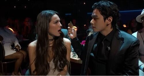 Kim Taehyung and Olivia Rodrigo sitting together at the 2022 Grammy's, Taehyung in a black suit with a card in his right hand, Olivia looking surprised